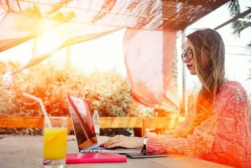 Woman in glasses working on a laptop outdoors at a sunny café with orange juice and a tablet on the