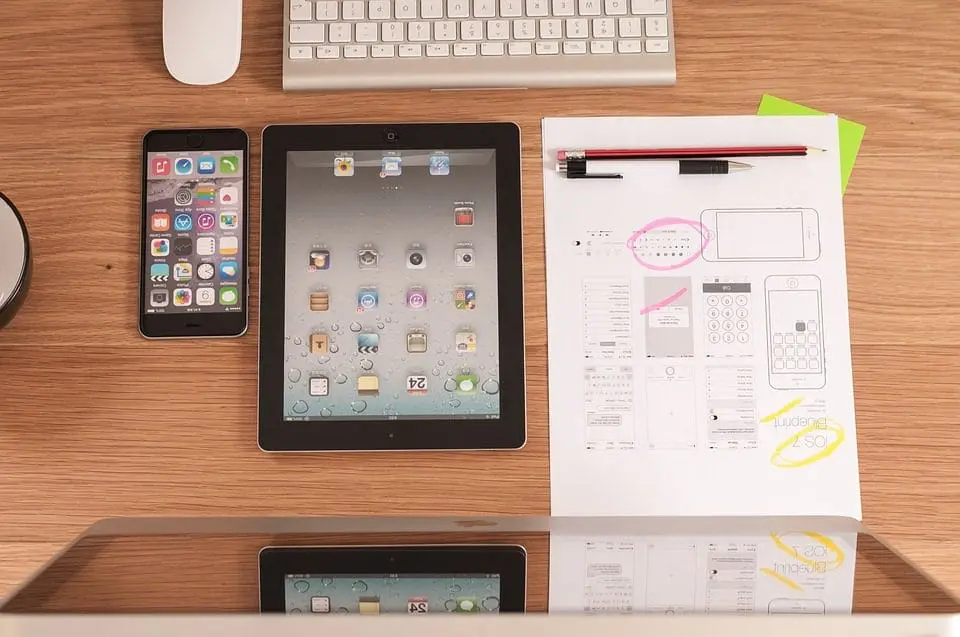Workspace with an iPhone, iPad, UX design sketches, pencil, and keyboard on a wooden desk