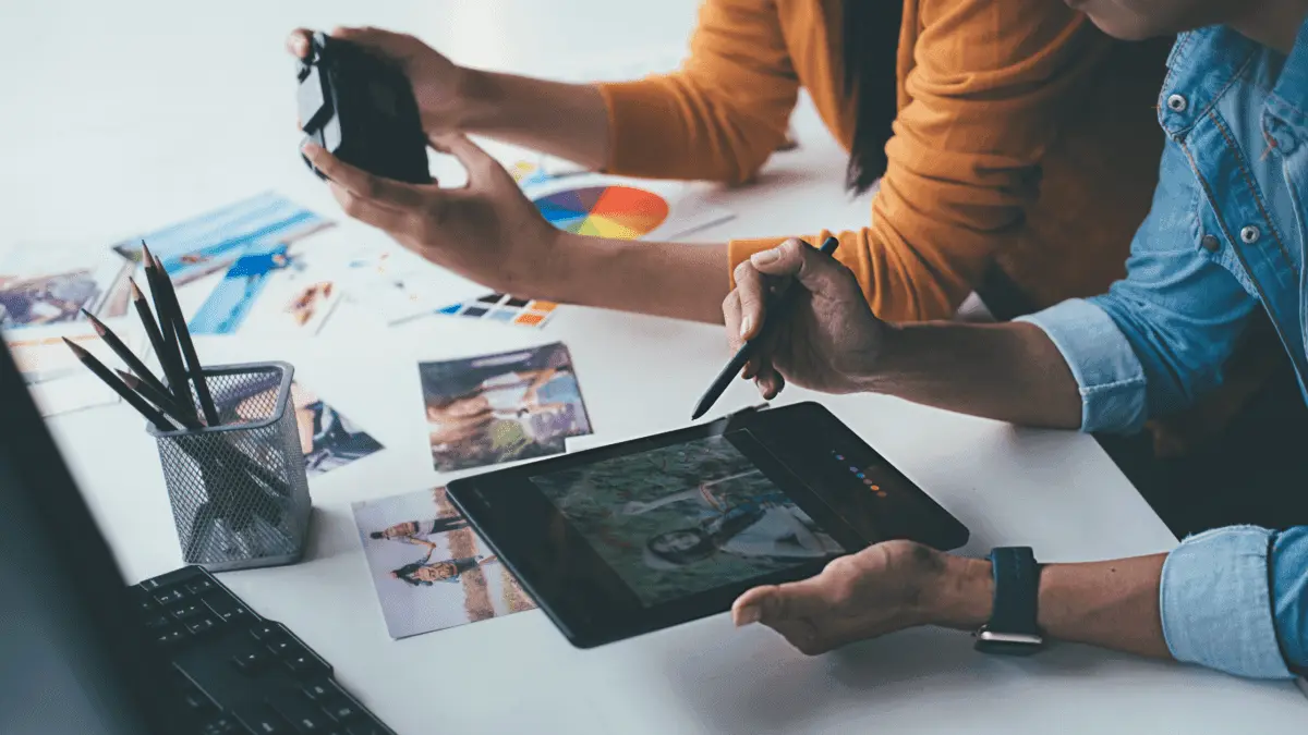 Two people collaborating on photo editing using a tablet and digital pen, surrounded by printed