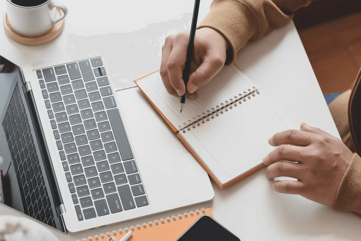 A person writing in a notebook with a laptop open in front of them.